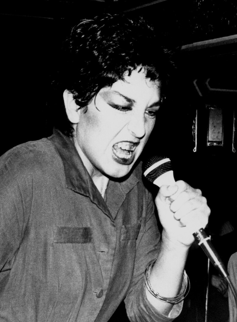 Singer Alice Bag holds a microphone, with a snarl on her face.