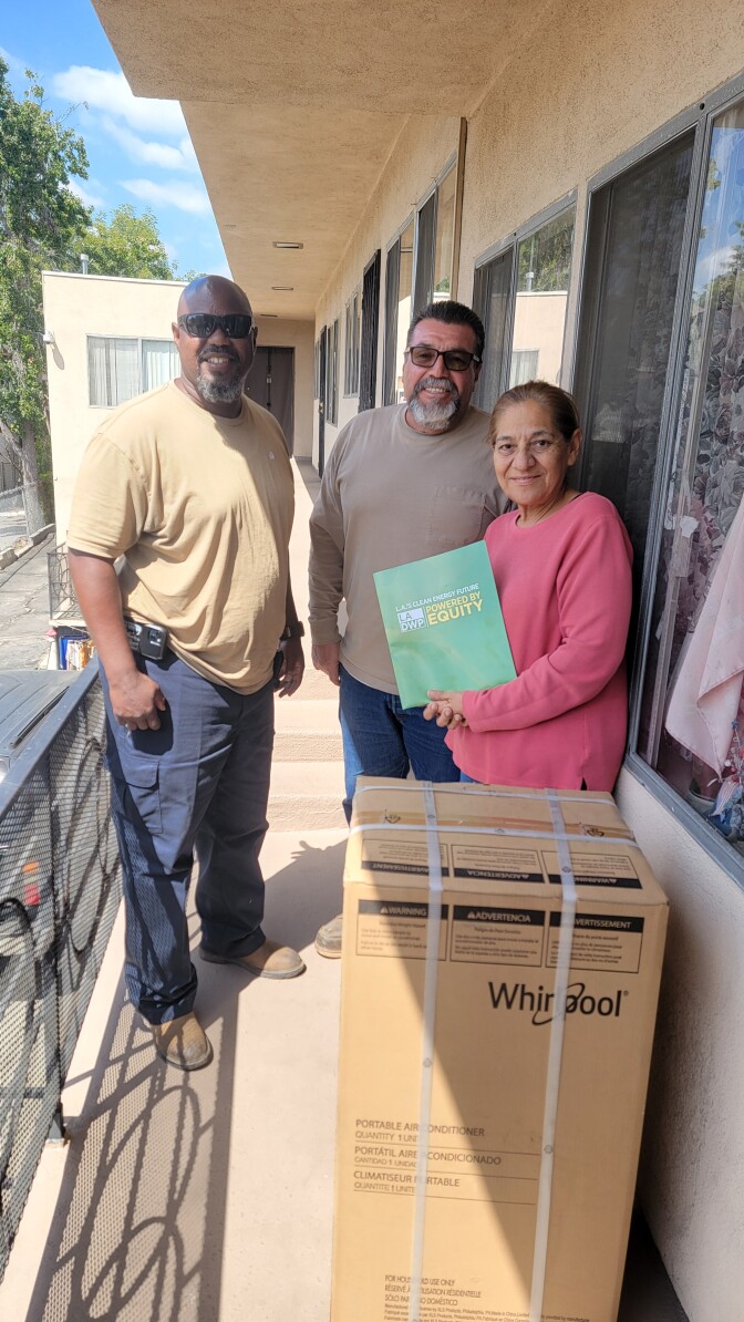 A middle-aged Black man wearing a khaki t-shirt and blue work pants and boots stands next to a middle-aged man with light brown skin and dark hair and greying beard and mustache wearing a khaki long-sleeved shirt and jeans standing next to an older Latina woman wearing a salmon pink long-sleeved shirt. A large box is in front of them and they stand on a narrow balcony. They all smile. It's sunny. 