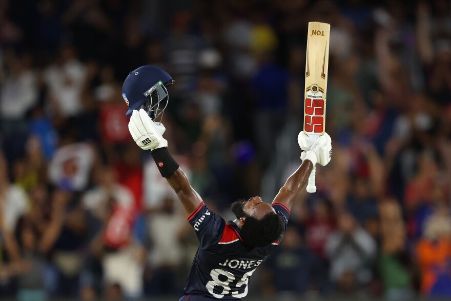 A man with both arms raised. In one hand he is holding a helmet. In another a cricket bat.
