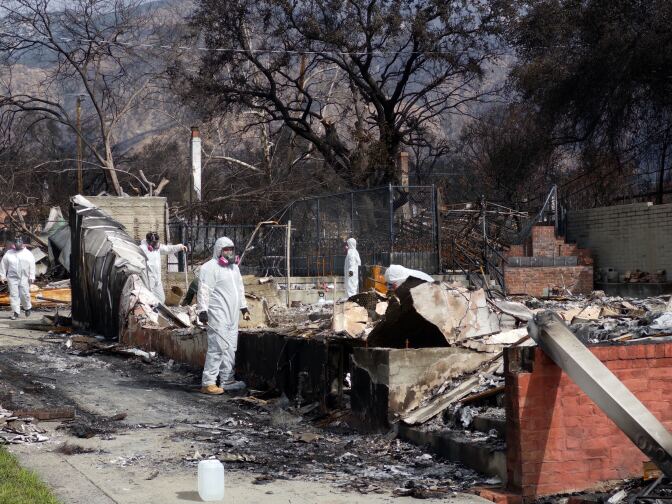 Workers in white suits covering their bodies and face masks remove debris from a burnt home.