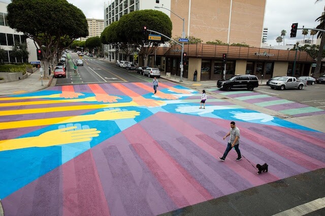 An ariel view of a crosswalk intersection with a colorful design on it, including bright yellow hands. People walk across while cars wait.
