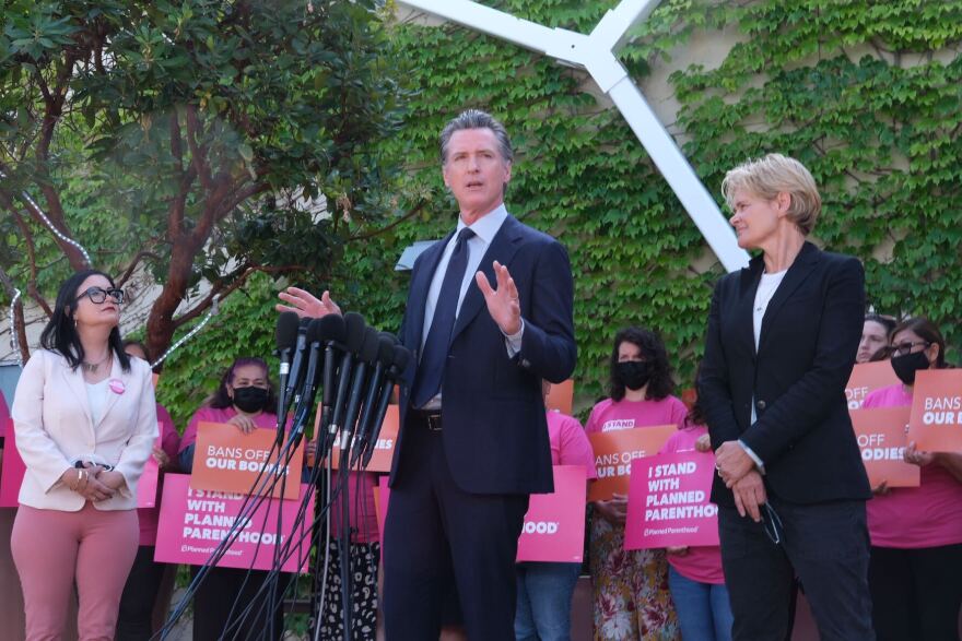 Governor Gavin Newsom, wearing a dark suit and tie, speaks before a cluster of microphones, flanked by a women on either side of him. Behind him are about a dozen people holding pinks signs that say "Bans off our bodies" and "I stand with Planned Parenthood."