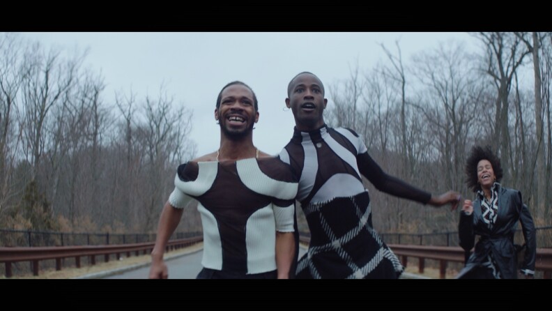 A view of two Black men wearing black and white abstract outfits in mid-movement on a road outside on a cloudy day. They appear excited, looking toward something in the distance. In the background is another Black person who's dancing.
