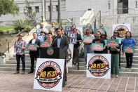 A man with short dark hair and medium light skin tone is dressed in a light blue collared shirt and sports coat, speaking at a podium. Behind him, about twenty people hold up signs that read "ICE out of L.A." and "Keep Families Together." They 