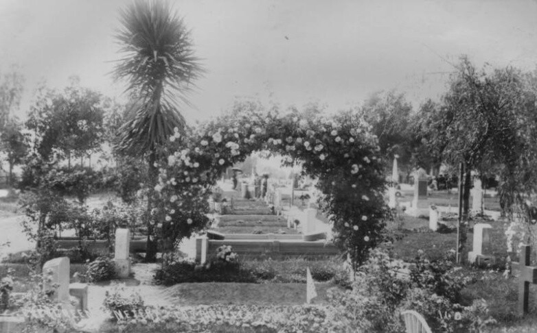 Faded black and white image has an arch of flowers amid grave stones.