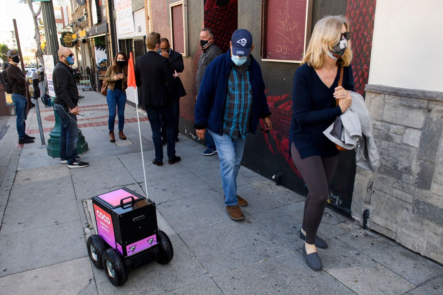 A small pink four-wheeled delivery robot rolls on a Los Angeles sidewalk alongside pedestrians.