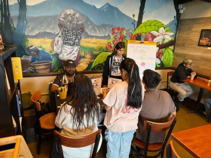 Two people behind a table in front of a mural on a wall listen to a small group of people speaking across from them. The table has signage and a large post it board stands next to them.