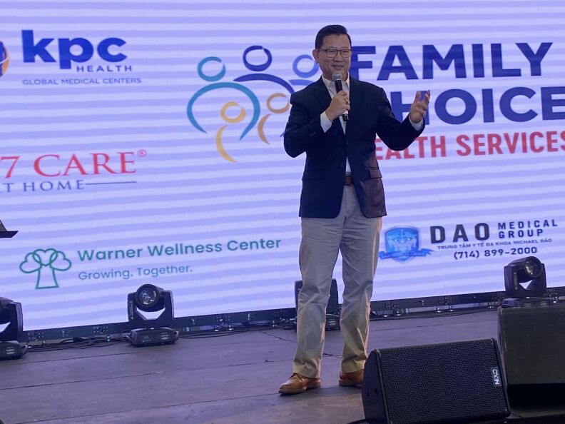 A man speaks on a stage into a microphone in front of a logo for Warner Wellness Center