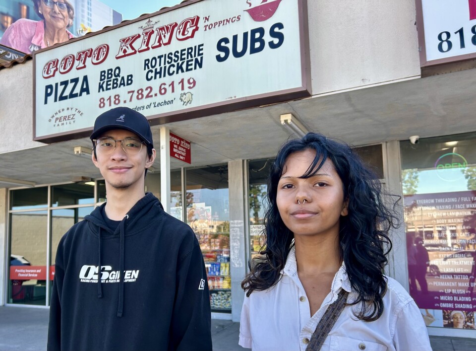 A person wearing a white shirt and a person wearing a dark sweatshirt stand in front of a pizza shop.