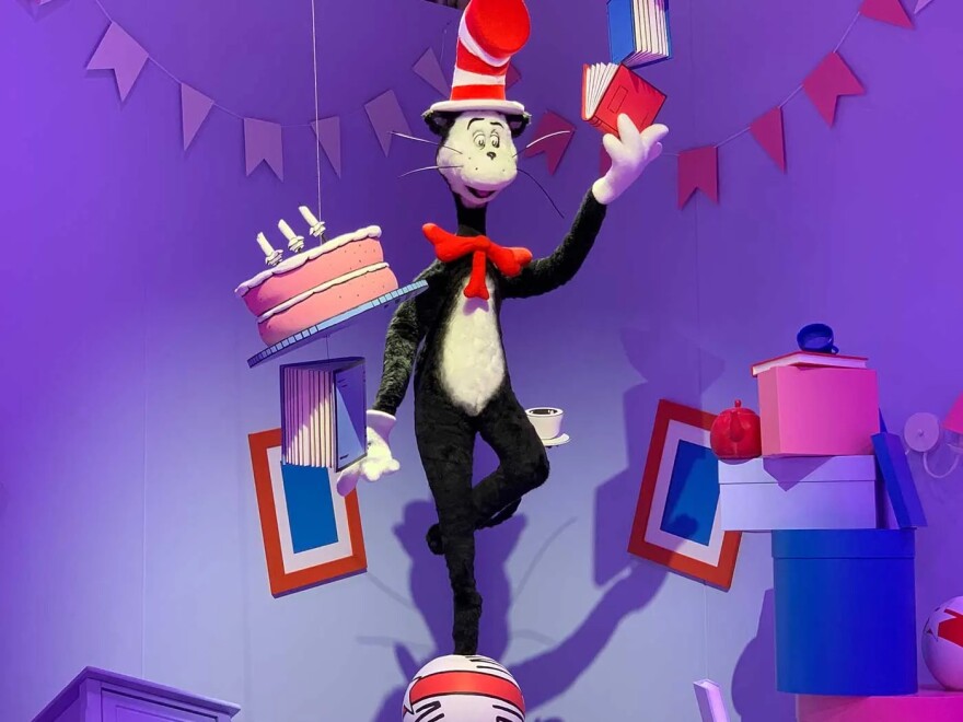 A giant puppet version of the Cat in the Hat character from Dr. Seuss books stands on a ball and holds a book in hand in a room full of streamers, boxes, with picture frames on the walls behind him. He is black with a white belly and is wearing a red and white-striped top hat.