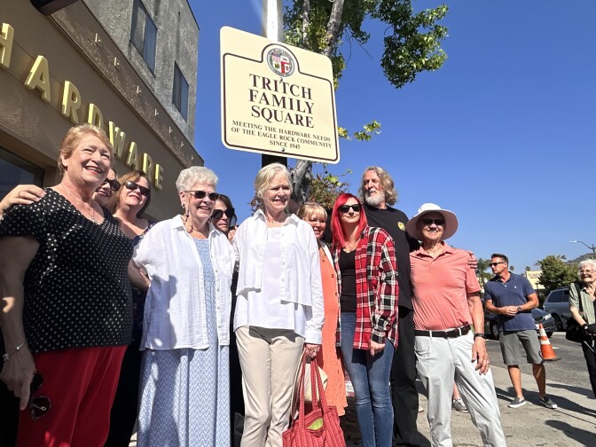 A crowd of people of varying ages are smiling and posing in front of a city street sign that reads "Tritch Family Square" and "Meeting the hardware needs of the Eagle Rock community since 1945" on a cloudless, sunny day.