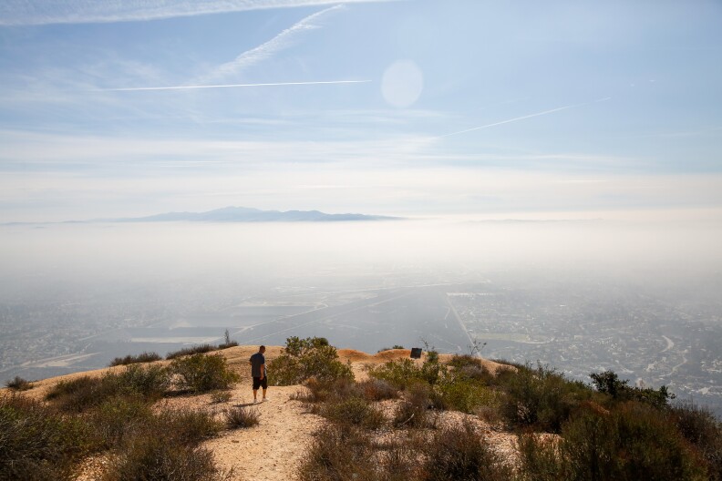 A photo of a man by the side of a cliff overviewing a community with a mountain range in the horizon.