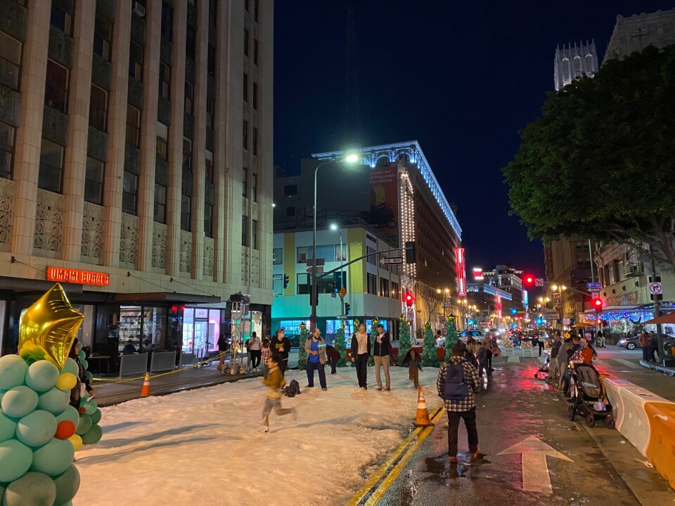 Kids play on a patch of snow at night on Broadway St. in downtown Los Angeles while parents watch.