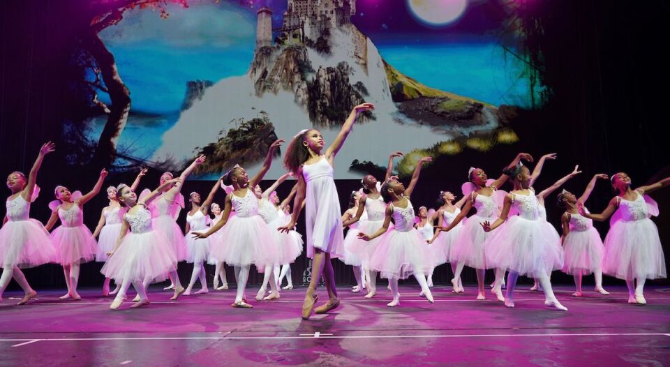 A number of young POC ballerinas dance on stage in white ballerina outfits.