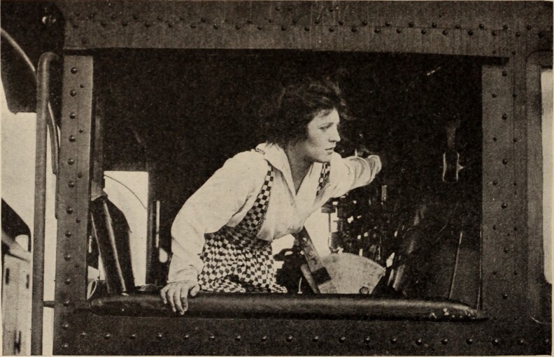 A black and white photo of a fair-skinned woman wearing a checkered jump suit and white collared shirt inside a locomotive car. Her head is turned toward the right as she drives the train.
