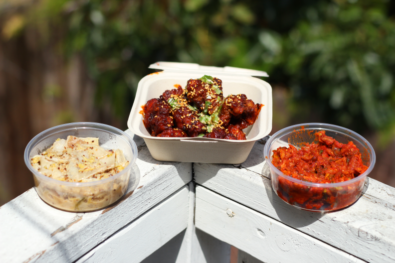 A cardboard container of fried chicken nuggets sits between two plastic containers of banchan on top of a wooden railing.