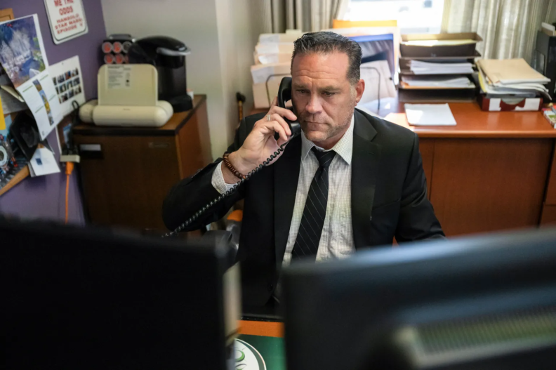 A man wearing a suit and tie sits at a desk holding a phone to his ear and looking at a computer monitor.