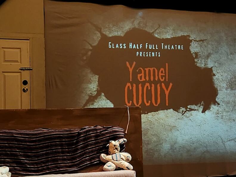 A stage dressed to look liek the inside of an apartment. There's also a projection screen displaying the name of the show, 'Yamel Cucuy."