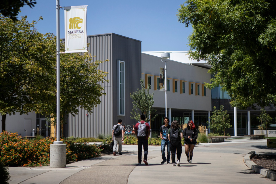 students wearing backpacks walk outside on their community college campus in front of a gray building with a yellow mascot on the side