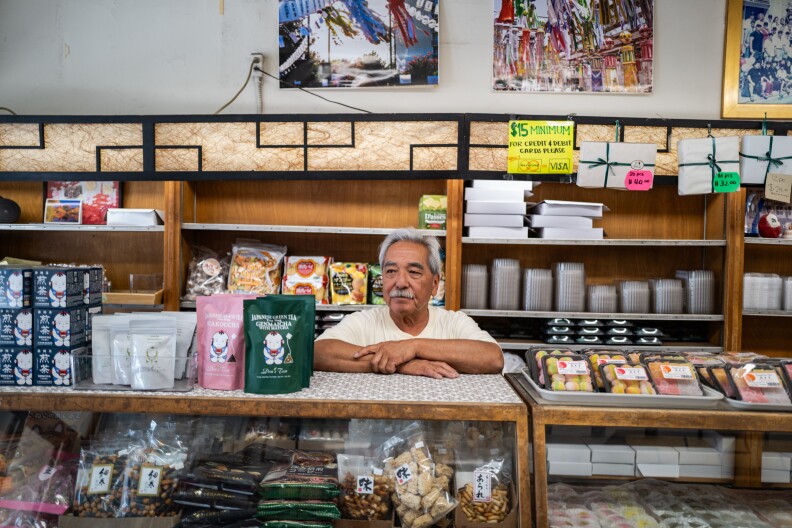 A medium skinned man with grey hair leans on an old fashion wooden shop counter. He's surrounded by colorful boxes and bags of sweets