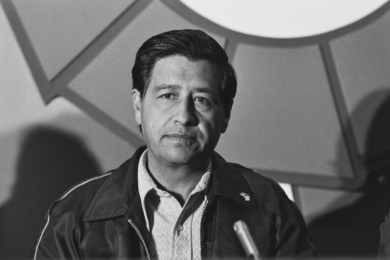 A black and white image of Cesar Chavez in a jacket and collared shirt.