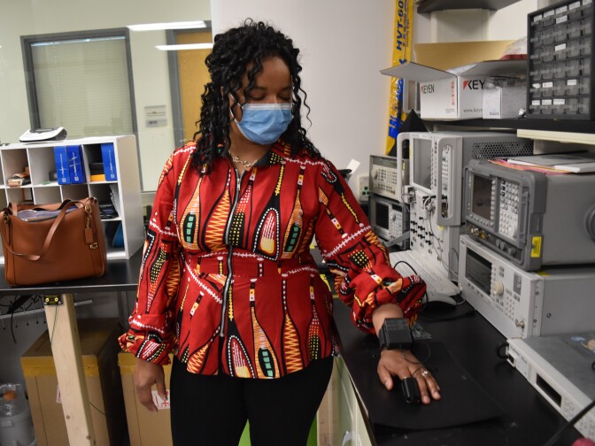 A Black woman wears a prototype for a new type of pulse oximeter her lab has patented. She stands at a counter in a lab. Several machines are on the counter.