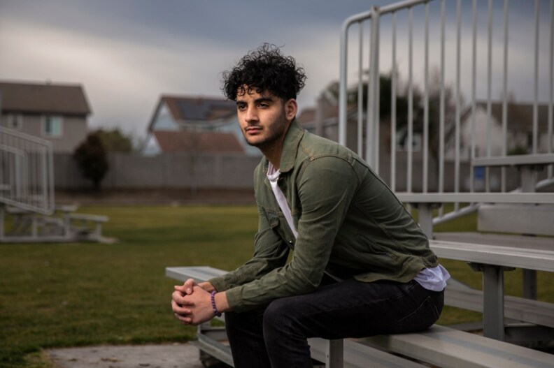A young man with Brown skin, scruffy facial hair and tousled curly black hair sits on bleachers in the middle of a green field. Houses can be seen behind him, which are behind a wooden fence. He wears a green jacket, white shirt and dark pants and looks into the corner of the image.