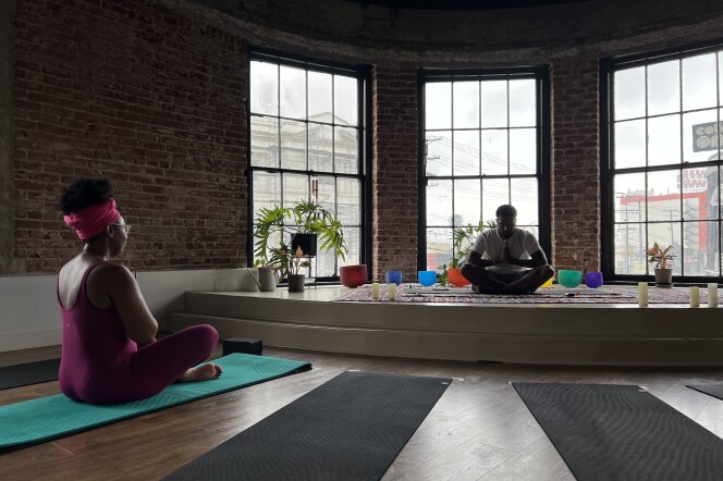 Two brown skinned people sit cross-legged in a dark studio room on yoga mats. 