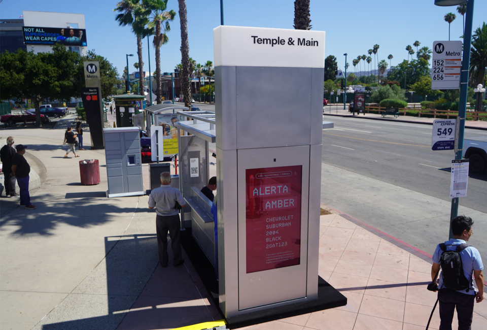 A person stands in the shade provided by a bus shelter as other people stand and walk around at a transit station next to a street.