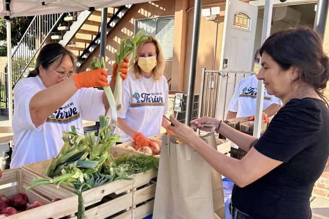 A female presenting person puts vegetables into a paper bag held by a female presenting person.