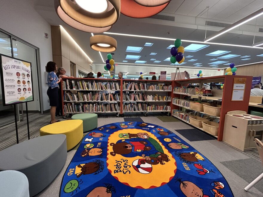 In the inner corner of a bookshelf, a colorful carpet with cartoon animals and kids under round and square lights. There are balloons around the room. A female-presenting person stands to the left of the shelf and more people's heads peek out from behind the shelf.