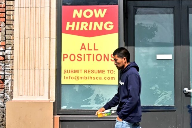 A man carrying a water bottle and wearing a blue hooded sweatshirt walks past a storefront with a sign hanging up that says, "Now hiring, all position" with information on where to send a resume.