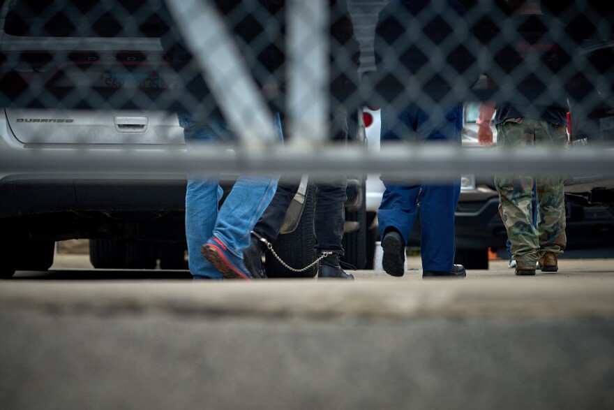 A low angle view showing the pants and shoes of people walking away towards cars, including one person who has chains on their legs. A gated fence is out of focus in the foreground.