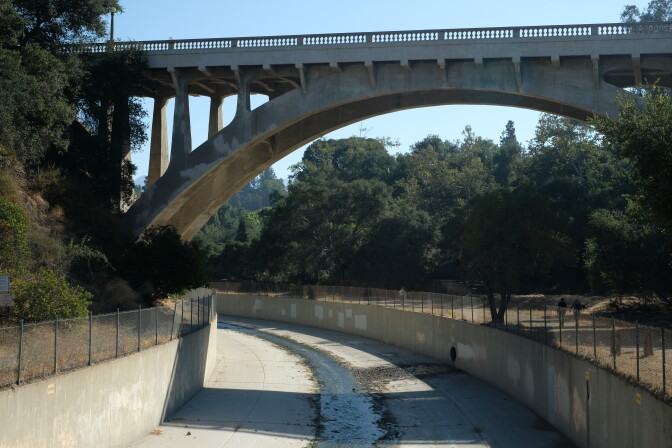 A concrete arched bridge goes over a channelized stream. Lush trees are on either side. 