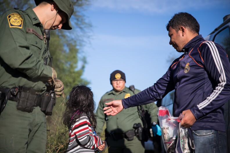 A young child with long hair looks at an ICE official. A man not in uniform stretches his hand out toward her.