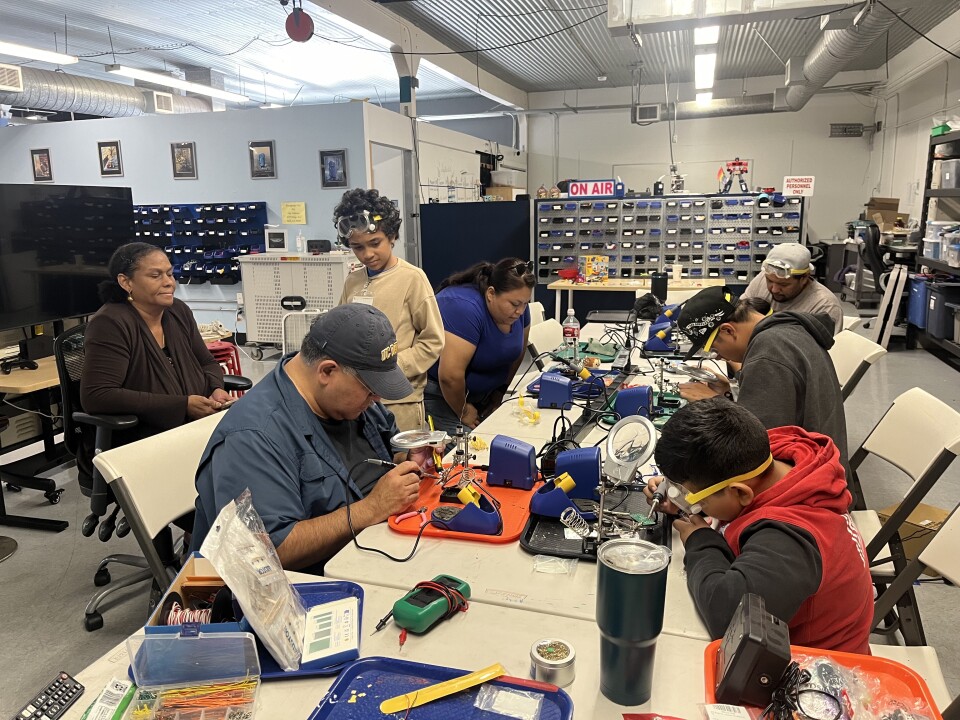 A group of kids with some adults sit around a table. The kids are wearing safety glasses and are concentrating on the electrical work they're doing.
