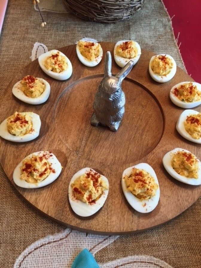 A group of deviled eggs are arranged in a circle on a wooden plate on a table. In the middle of the plate is a decorative metal rabbit