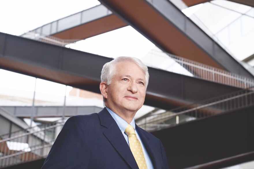 Man with gray hair and suit and tie looks into the distance, behind him stairways