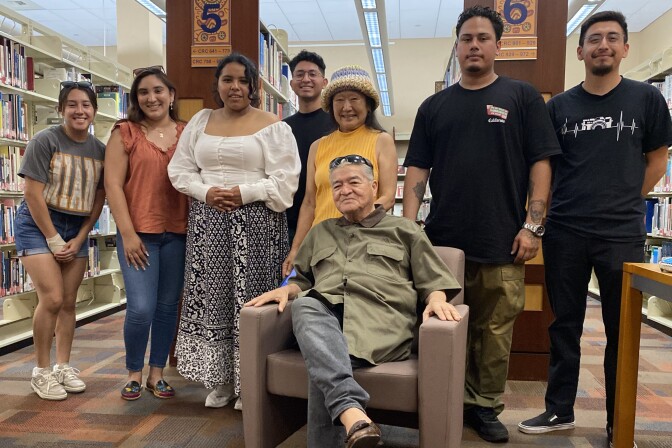 Josof Sanchez (seated, center) invited a group of Latino youth to open up about their mental health journey in order to help others. They are in a library surrounded by book shelves. 