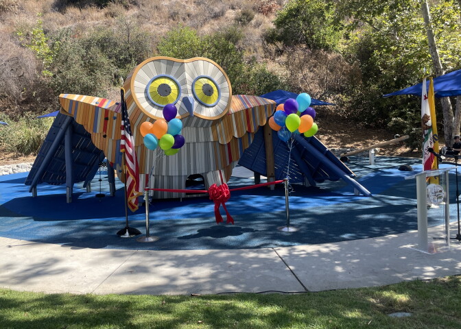 A 12-foot owl-shaped playground is seen on a rubber mat surface, surrounded by grass, balloons and vegetation.