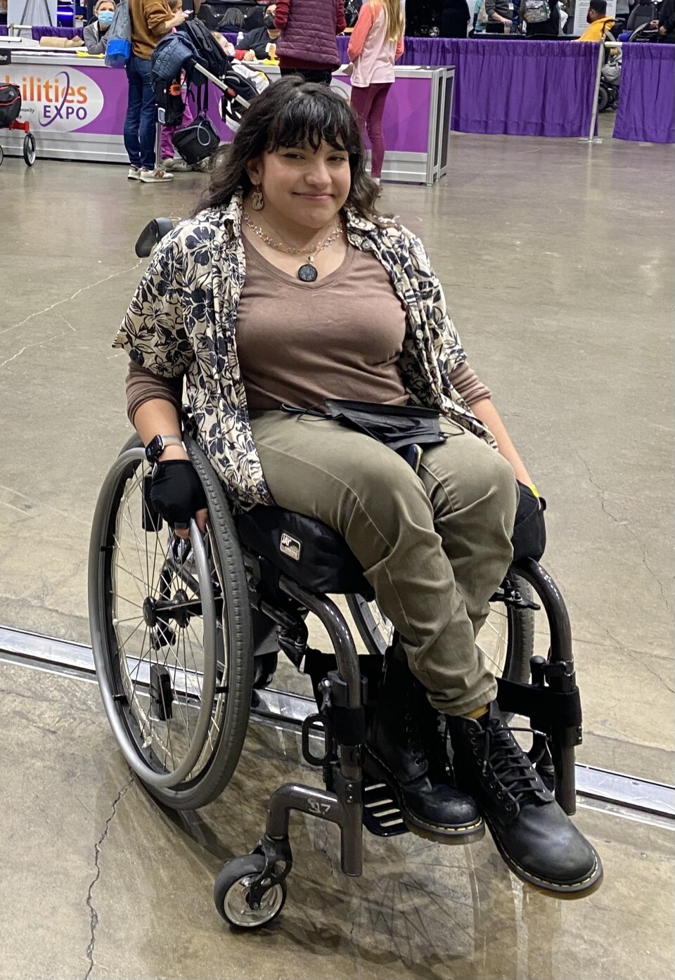 A woman with long dark hair, black boots and olive green pants poses in a wheelchair, smiling, in the middle of a large room.
