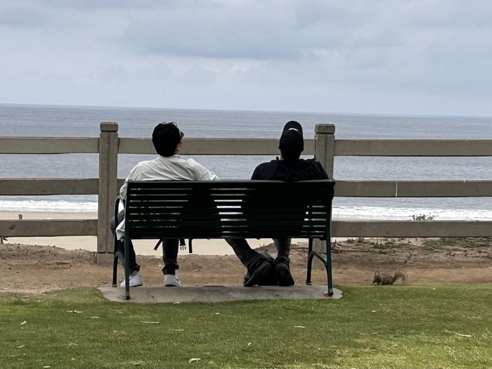 Two men are sitting on a metal park bench looking out towards the ocean on a hazy, cloudy day. A small brown ground squirrel is sitting off to the right hand side.