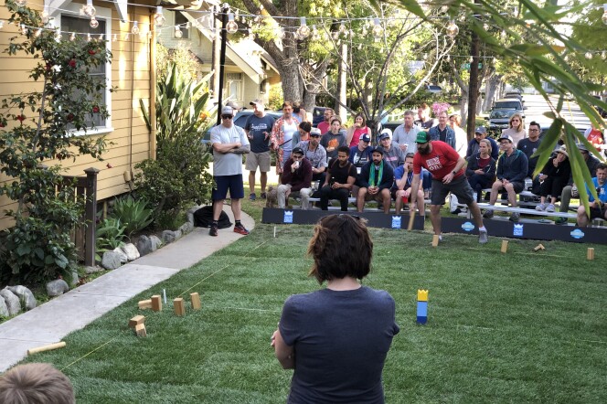 A group of people congregating around a backyard lawn. The lawn has wood blocks lined up on it, as a man in a red shirt tries to knock them down by tossing a baton.