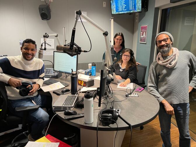 Four people gather around a desk in a radio studio, from left a man with dark-tone skin wears a white sweater with a strip, two women with light-tone skin and a man with dark-tone skin wearing a scarf and cap 
