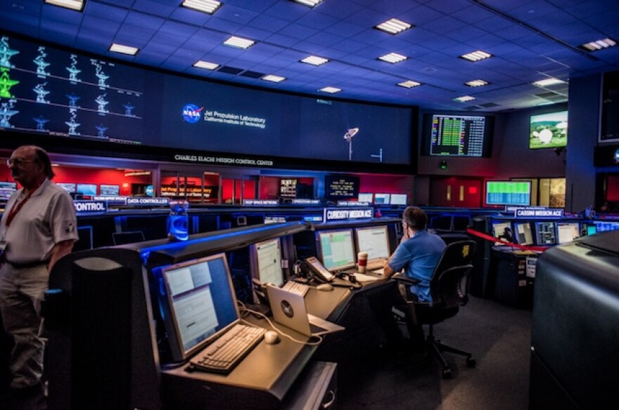 A NASA/JPL employee sits in front of a row of computer screens while looking up at a wall of monitors inside JPL's Control Center.