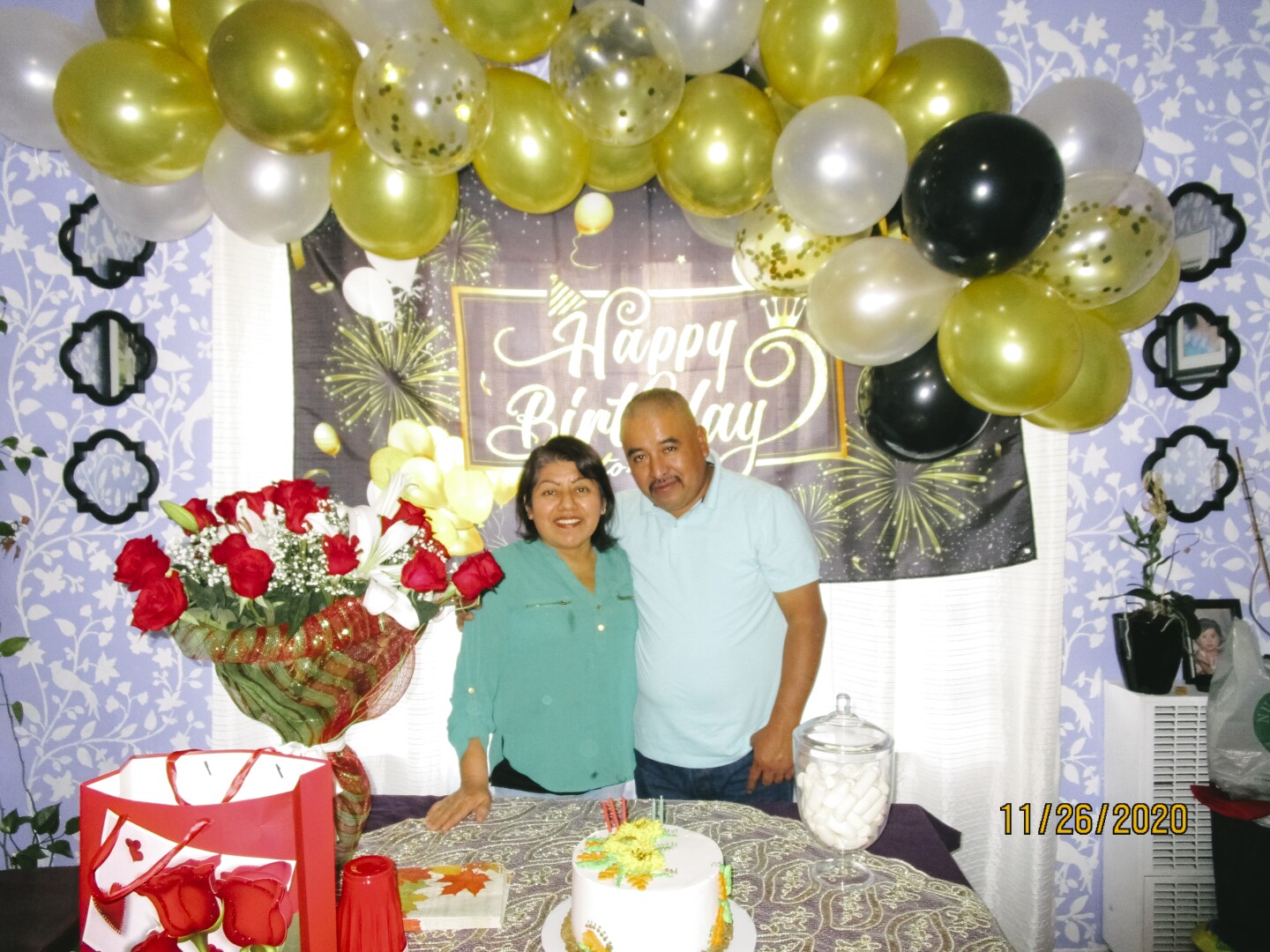 This is an image of Luz Hernandez and her husband. The walls are decorated for Luz's birthday. Gold, black, and white balloons hang on the wall. A cake and a bouquet of flowers are on the table.
