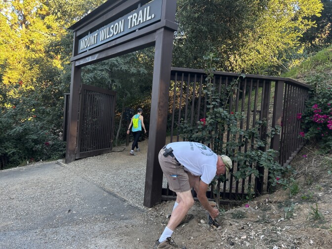 A woman with a neon backpack and exercise clothes walks under a large metal sign across a trail that reads "Mt. Wilson Trail." To the right an older white man bends over doing some work beside the trail. 