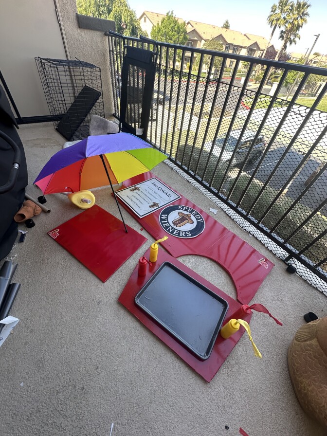 An apartment patio overlooking a parking lot with three red pieces of a soapbox laying on the ground. There's a rainbow striped umbrella set up to the left of the pieces, with a yellow mustard bottle and red ketchup bottle affixed to the right with matching streamers hanging from the nozzles.
