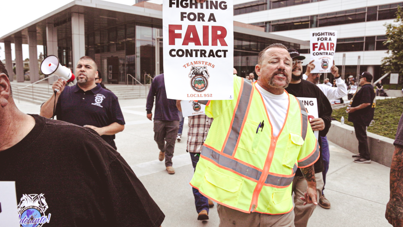 A man in a neon vest is walking and holding a sign that reads "For a Fair Contract Teamsters Local 952". Crowds of people behind him are also walking and holding signs.