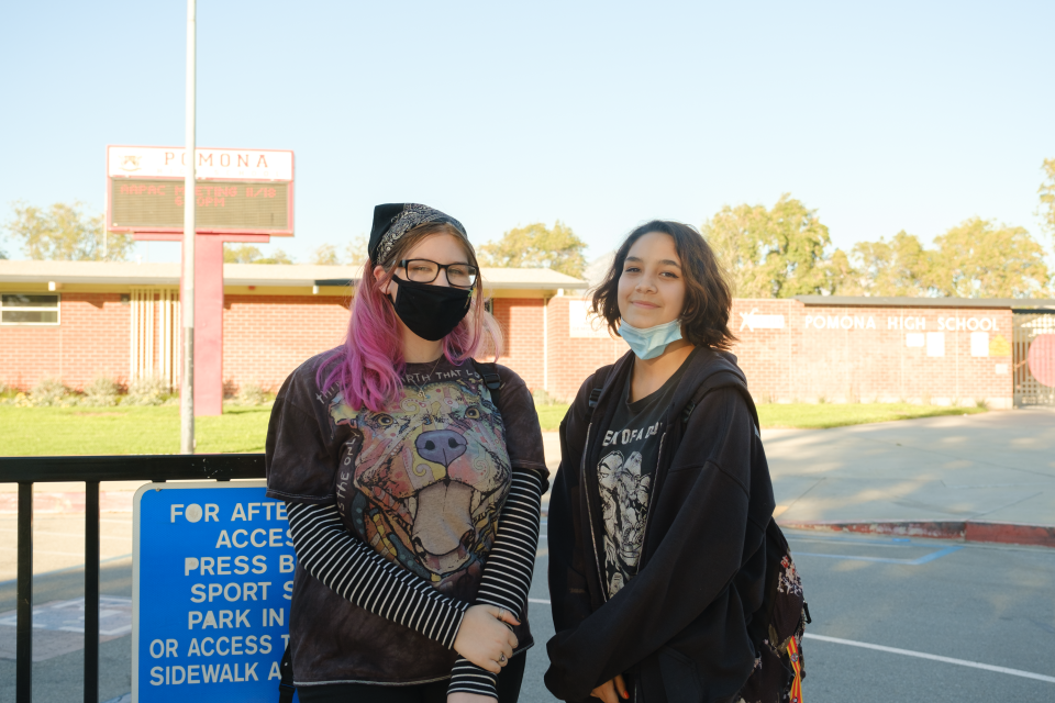 Two young girls pose for the camera in front of a building with a sign that reads "Pomona High School." The girl on the left has pink hair, wears a mask, and a bandana. The girl on the right has her mask pulled down and smiles.